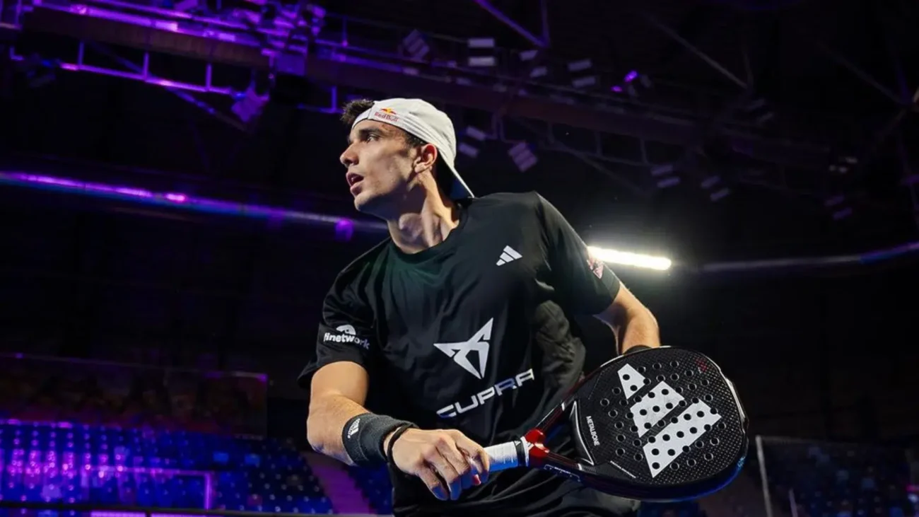 Eine Person in schwarzem Sportoutfit und weißer Kappe spielt Paddle-Tennis in der Halle. Sie möchte wahrscheinlich ihre Position auf der offiziellen Rangliste der Padelspieler FIP verbessern. In einer Hand hält sie einen Schläger und konzentriert sich vor dem Hintergrund eines Netzes und einer violett beleuchteten Arena intensiv auf den Ball.