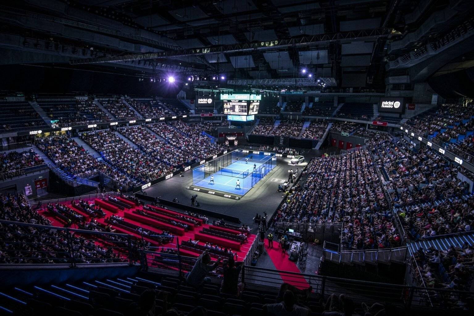 In einem voll besetzten Indoor-Stadion herrscht reges Treiben, während die Zuschauer ein Padel-Match auf einem hell erleuchteten Platz verfolgen. Große Bildschirme übertragen das Spiel und rote Teppiche säumen den Sitzbereich. Die Arena ist schwach beleuchtet, was die lebendige Atmosphäre dieses Events im Premier Padel Kalender 2024 perfekt unterstreicht.