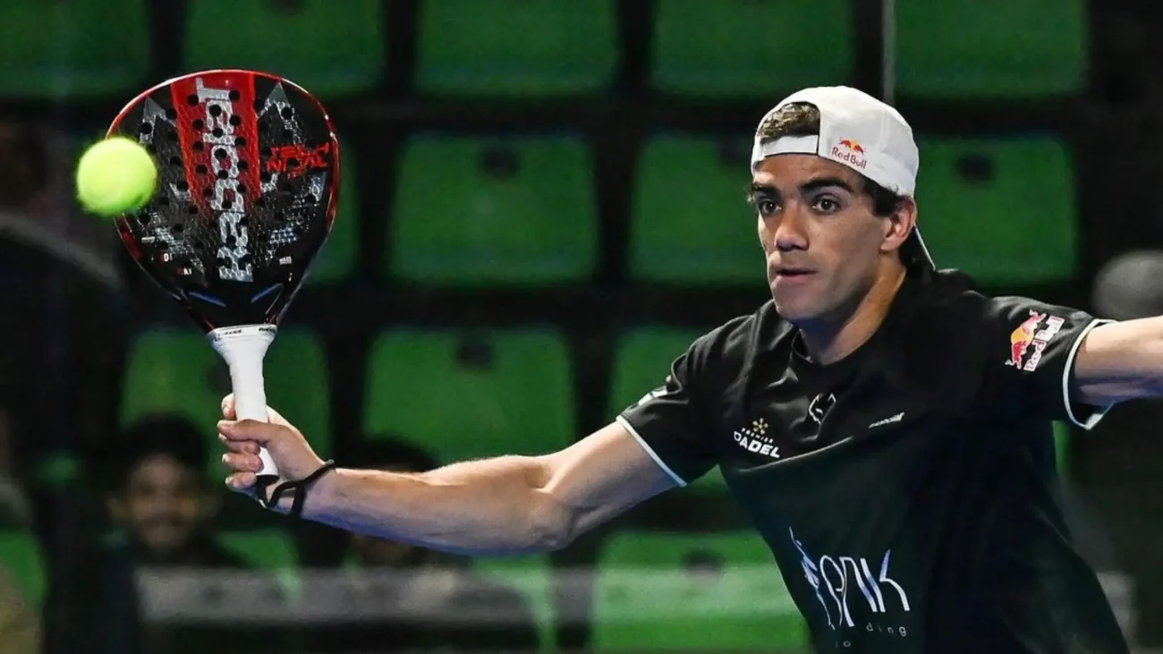 Ein Padelspieler in dunklem Sportoutfit und weißer Kappe, gelistet in der Offiziellen Rangliste der Padelspieler FIP, schwingt einen Schläger, um einen gelben Ball zu schlagen. Die grünen Sitze im Hintergrund lassen auf eine Halle schließen.