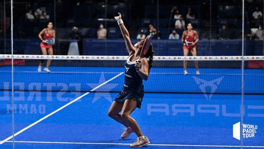 Ein Padel-Spieler im blauen Outfit bereitet sich darauf vor, den Ball mit einem Rückhandschlag zu schlagen. Im Hintergrund sind zwei weitere Spieler auf dem blauen Platz zu sehen, wo man sich fragt: „Was macht ein Profispieler nach einem Padelspiel?“ Das Logo der World Padel Tour ist auf dem Platz deutlich zu sehen.